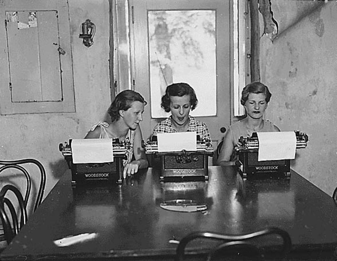 Three women sit at a table, each typing on a Woodstock typewriter. They are focused on their tasks, with sheets of paper in their machines. The room appears old, with worn walls and simple furniture.