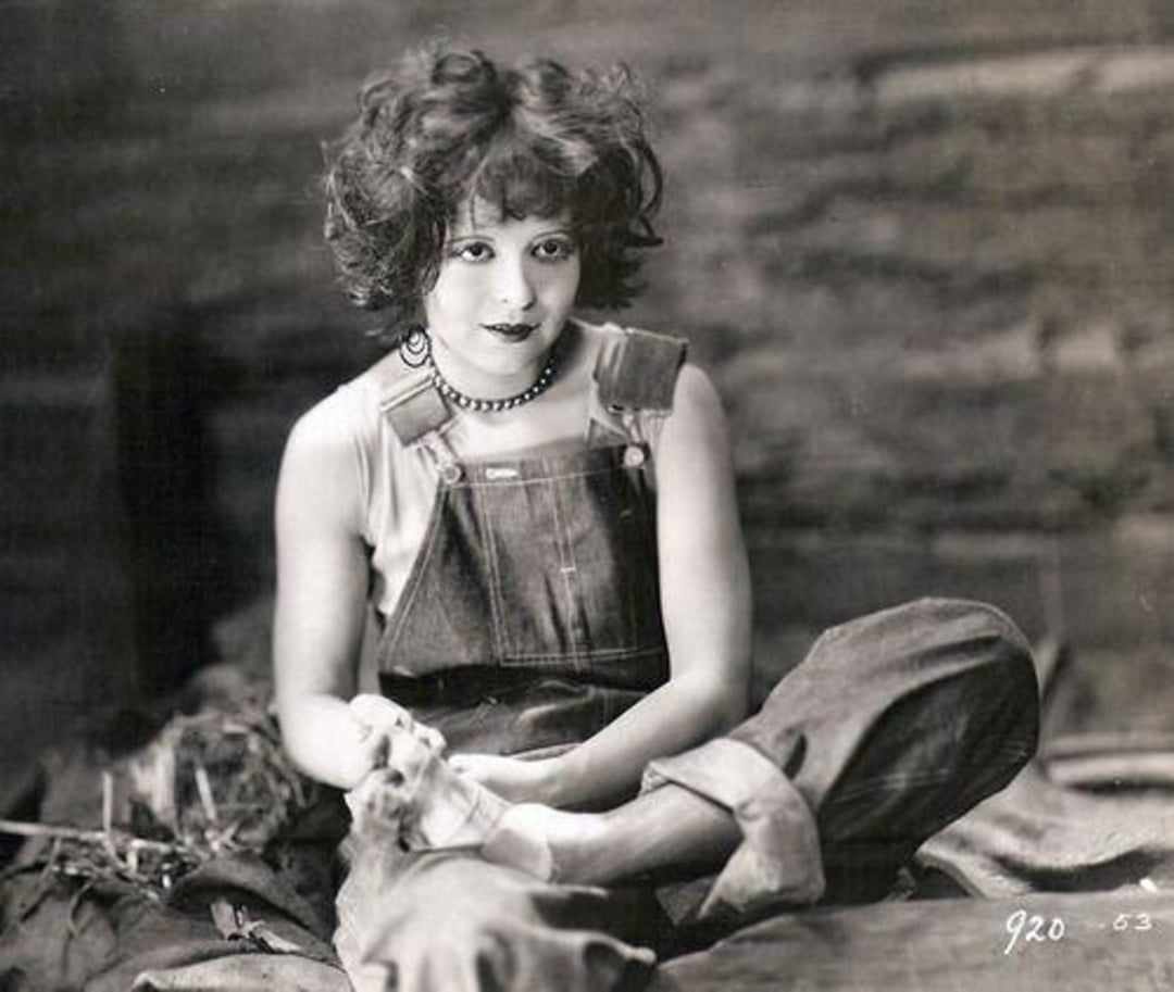 A young woman with curly hair, wearing overalls and a sleeveless shirt, sits cross-legged on the ground, looking at the camera with a slight smile. She holds a cloth in her hands. The photo is in black and white.