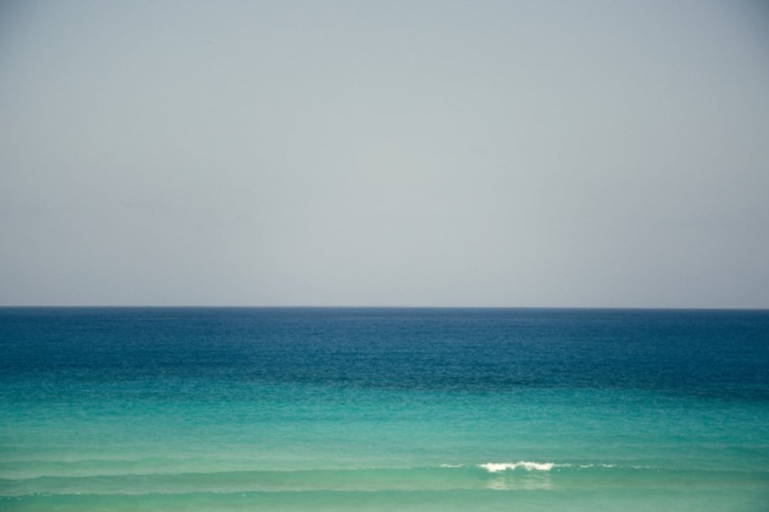 A calm, turquoise ocean stretches to the horizon under a pale blue sky, with gentle waves and no visible land or objects.