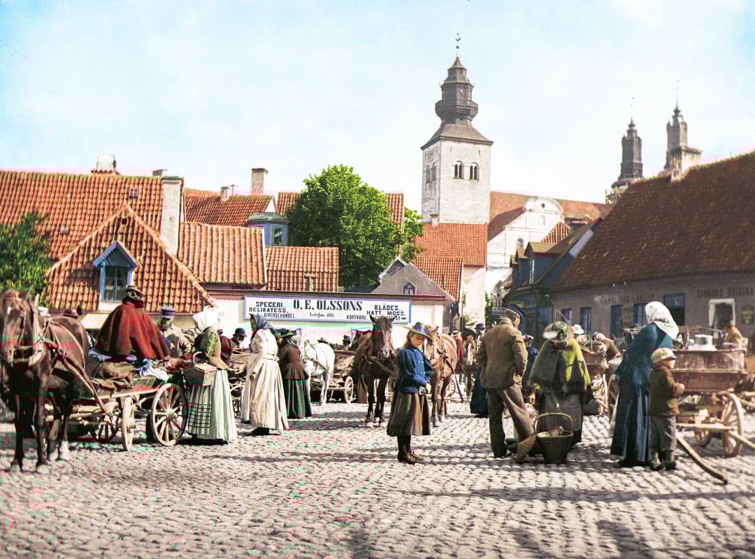 A bustling old town market scene with people in historical clothing, horse-drawn carts, and cobblestone streets, set against red-roofed buildings and a church tower in the background.