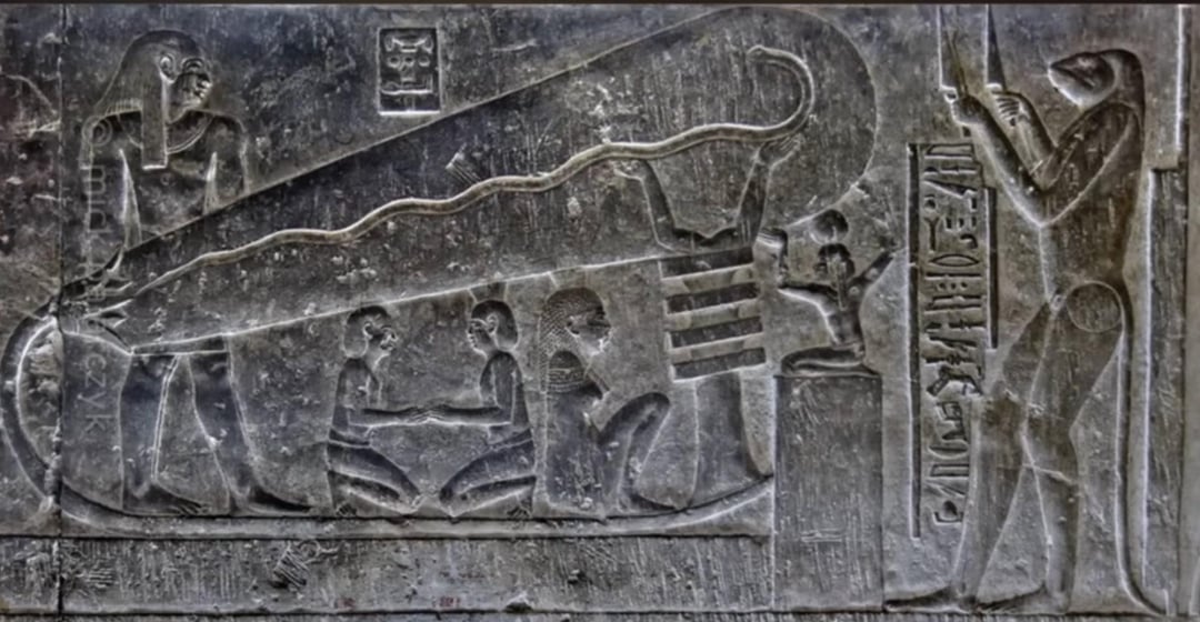 Ancient Egyptian stone carving depicting three seated figures inside a large oval shape with a serpent, a standing figure on each side, and hieroglyphs carved alongside the scene.