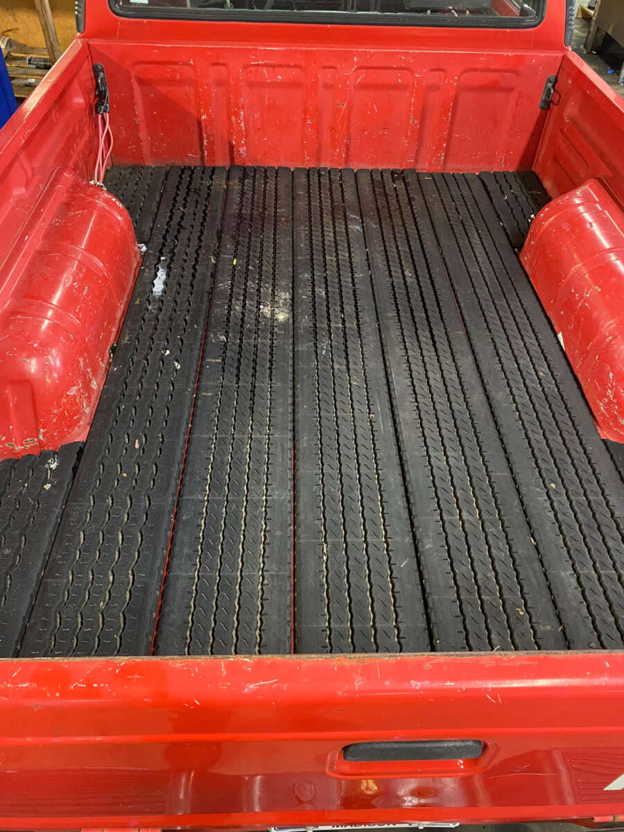 The image shows the bed of a red pickup truck with a black ribbed rubber mat. The truck bed has some visible scratches and scuff marks, and the mat features a tire tread pattern.
