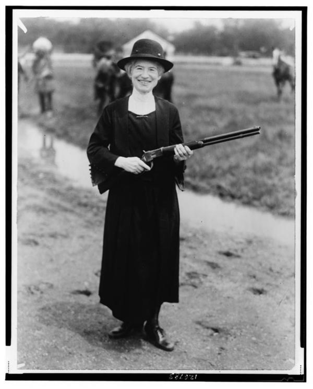 A smiling woman in a long dark dress and hat stands outdoors on a dirt path holding a shotgun. Blurred figures and grassy area are visible in the background. Image is black and white.