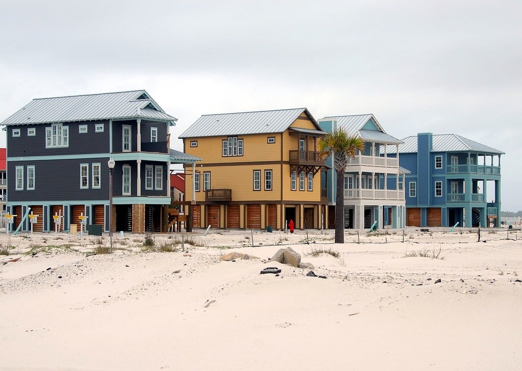 Colorful beach houses elevated on stilts stand in a row on a sandy, sparse beach under a cloudy sky. A lone palm tree and a few people are visible near the homes.