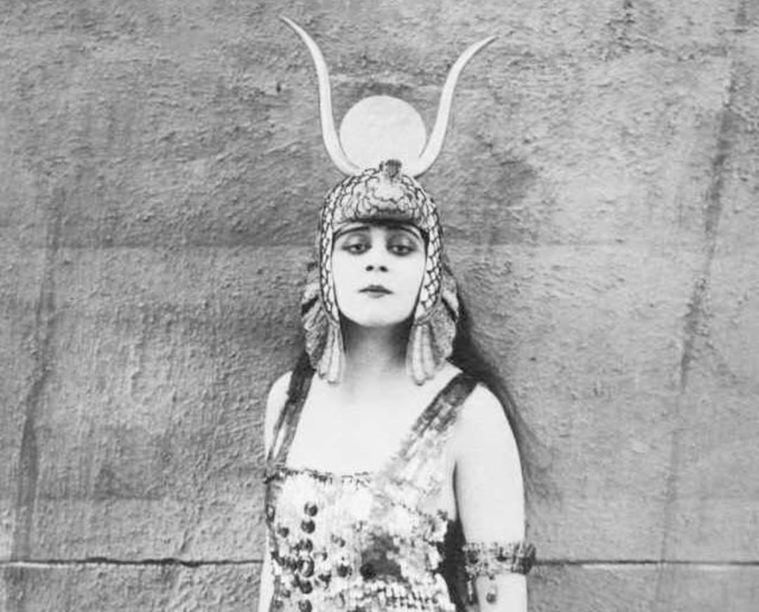 A woman in a sequined costume stands against a plain wall, wearing an ornate headdress with horns and a circular emblem, evoking an ancient or mythological style.