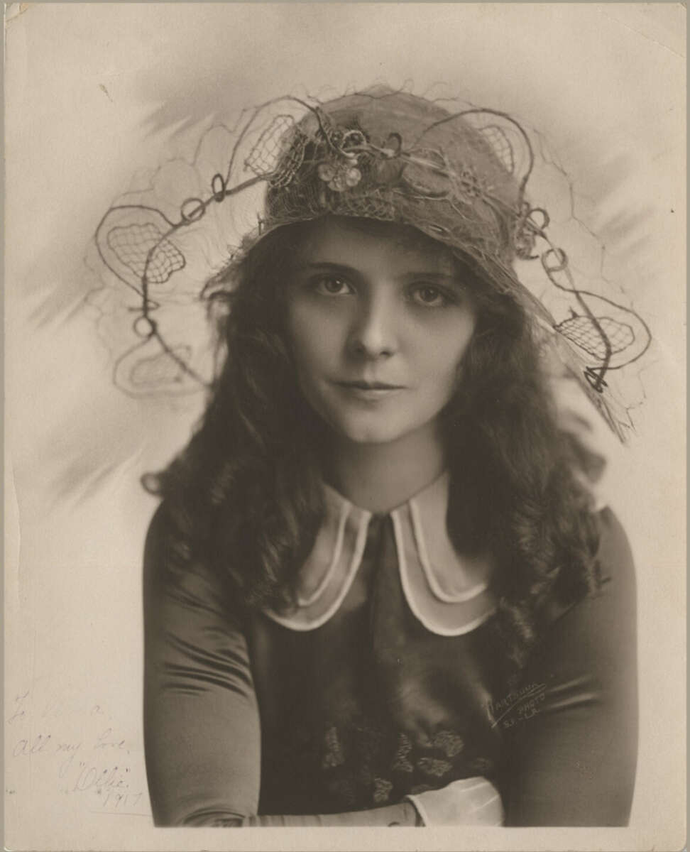 A young woman with wavy dark hair wears a wide-brimmed hat adorned with lace and decorative details. She is dressed in a dark dress with a white collar and gazes softly at the camera in this sepia-toned vintage portrait.