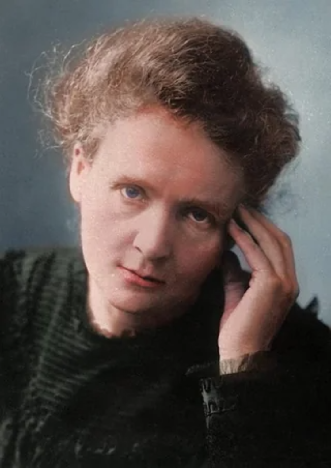 A woman with light skin and wavy, light brown hair rests her head on her hand, looking thoughtfully at the camera. She is wearing a dark, textured top and has a calm, serious expression. The background is softly blurred.