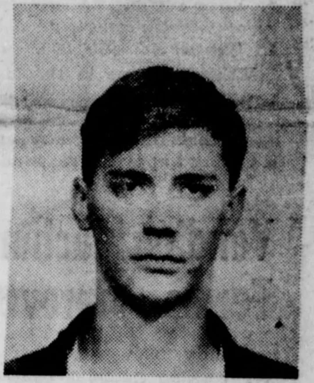 Black and white photograph of a young man with short, dark hair, looking directly at the camera with a neutral expression. The background appears plain or lightly textured.