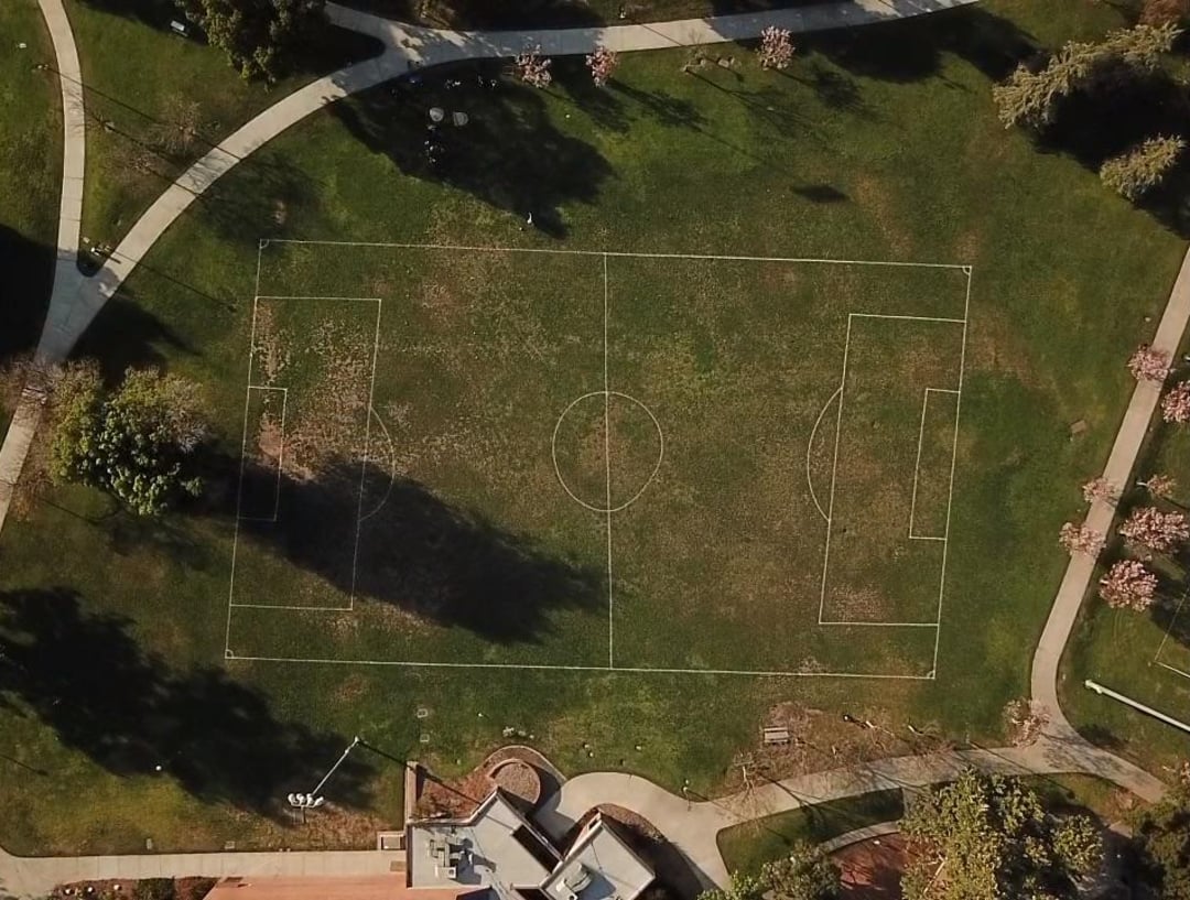 Aerial view of a soccer field with faint white boundary lines, surrounded by green grass, trees, walking paths, and a nearby building at the bottom edge of the image.