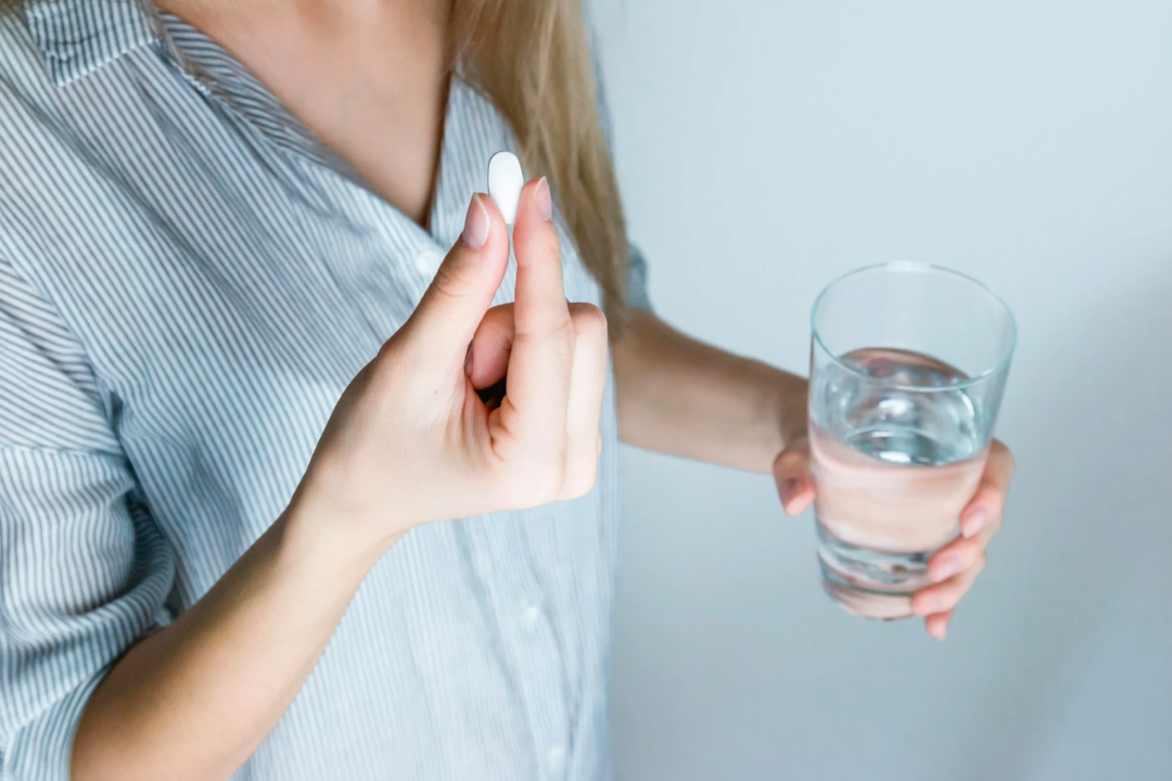 A person wearing a striped shirt holds a white pill between their fingers in one hand and a glass of water in the other, preparing to take medication.