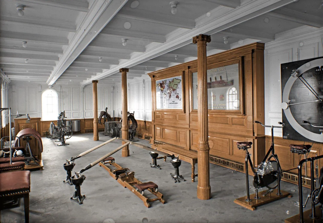 Vintage gymnasium room with wooden paneling, rowing machines, exercise bikes, and various fitness equipment arranged on the floor; maps and diagrams hang on the walls, and sunlight streams through arched windows.