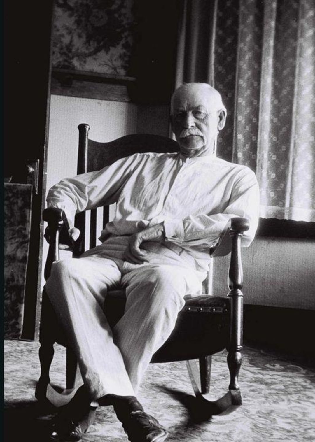 An elderly man with a mustache sits in a wooden rocking chair by a window, dressed in light-colored clothing, with his legs crossed and hands resting on his lap in a softly lit room.