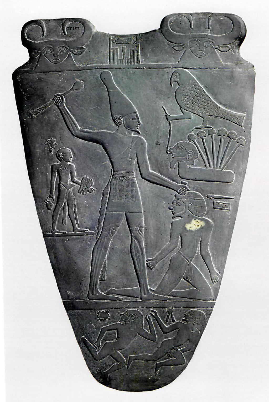 An ancient Egyptian stone palette showing a pharaoh holding a mace above a kneeling captive, with hieroglyphs and symbolic figures, including a falcon, carved around them.