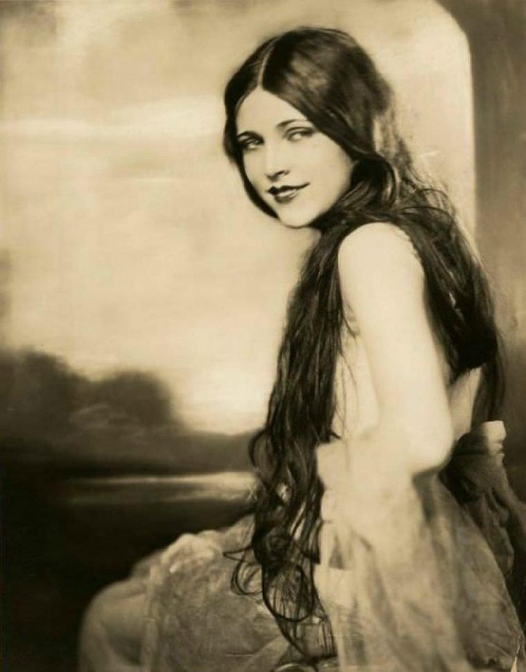 A vintage sepia-toned portrait of a woman with long dark hair, wearing a sheer, flowing gown, seated and looking over her shoulder with a soft smile in front of a blurred outdoor background.