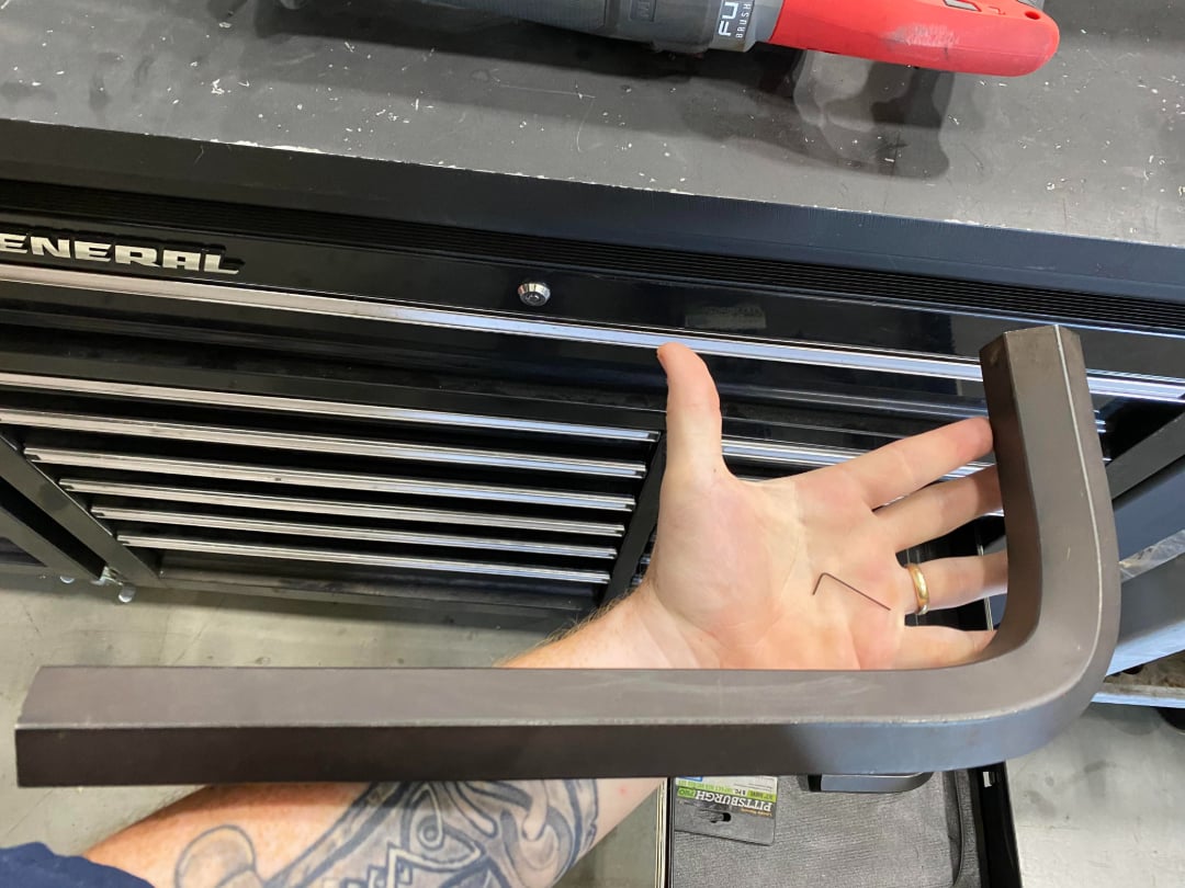 A person holds a very large L-shaped hex key in one hand and a small standard-sized hex key in the other, over a tool chest with drawers. A tattoo is visible on the person's arm.