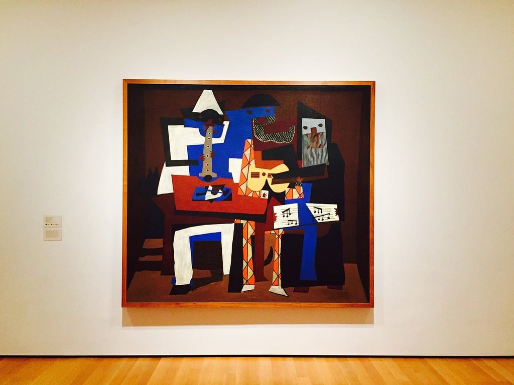 A large abstract painting in a wooden frame hangs on a white gallery wall. The artwork features three geometric, colorful figures playing musical instruments in a cubist style, set against a dark background.