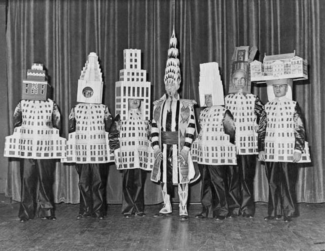 Seven people stand on stage in costumes; six wear outfits resembling New York skyscrapers, while the center person wears an elaborate costume and headdress. A curtain hangs in the background.