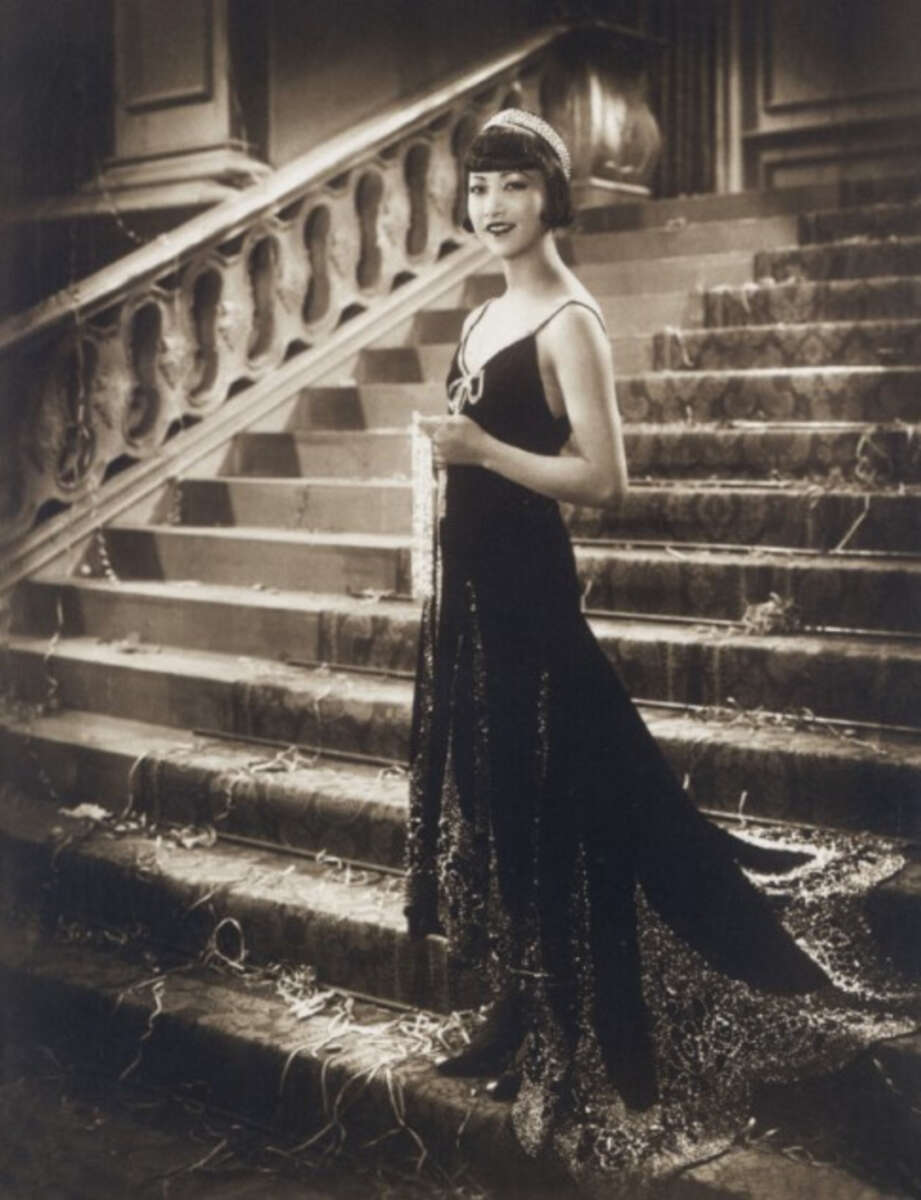 A woman in a black evening gown and headband stands smiling on a grand staircase, holding a clutch. The scene appears elegant and vintage, with confetti or streamers on the steps, suggesting a festive event.