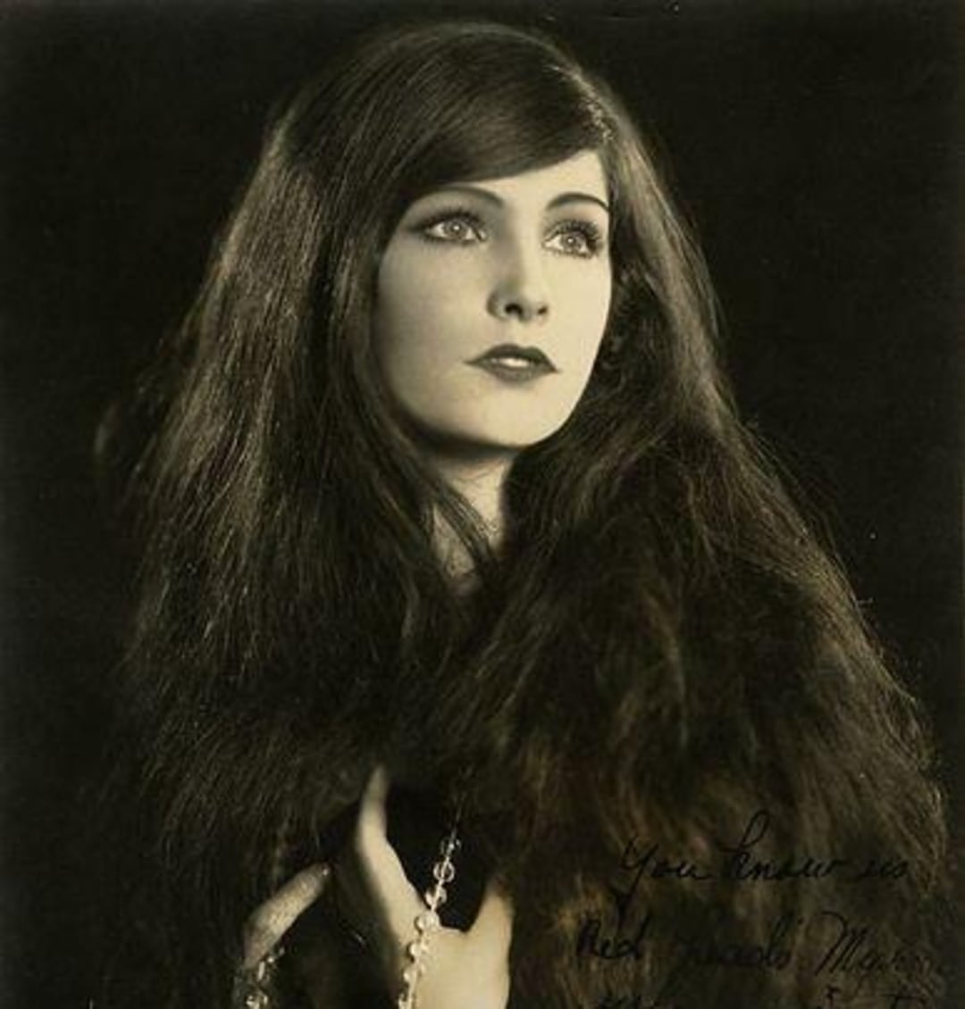 A black and white portrait of a woman with long, wavy hair, gazing slightly upward. She wears a fur garment and holds a necklace, with elegant makeup and a calm, thoughtful expression. Handwritten text appears in one corner.