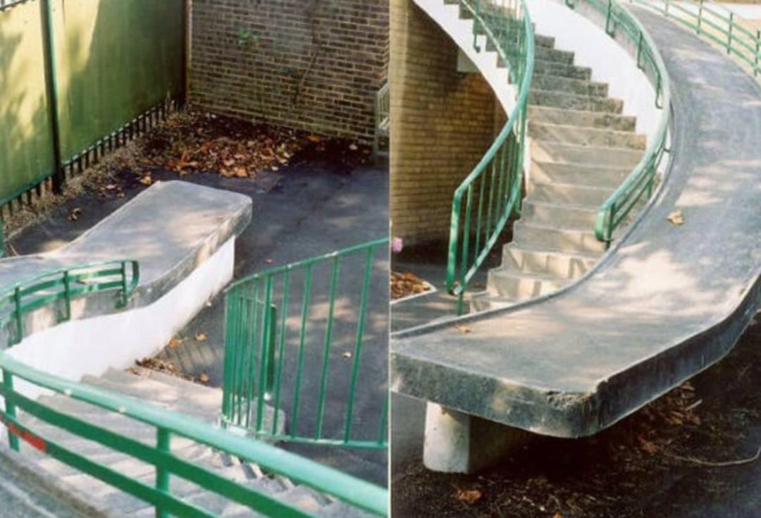 A concrete staircase with green railings, but instead of stairs on one side, there is a smooth ramp-like surface curving downward, creating an unusable or confusing design. Dry leaves are scattered nearby.