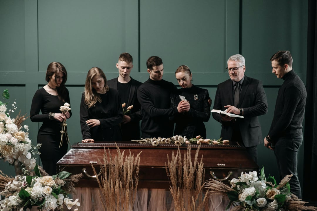 A group of people dressed in black stand around a wooden casket adorned with flowers, mourning at a funeral. One man holds a book, appearing to speak or read, while others look somber and hold flowers.