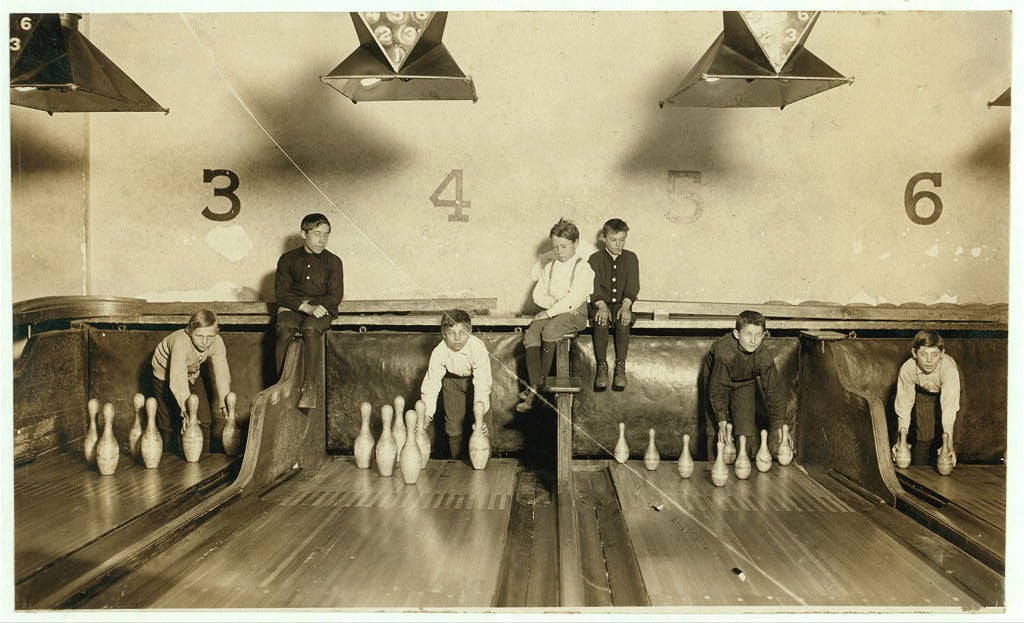 Seven children sit or kneel at the end of bowling lanes, each setting up pins by hand. The lanes are numbered 3 to 6 on the wall behind them. The image appears old, and the children are wearing vintage clothing.