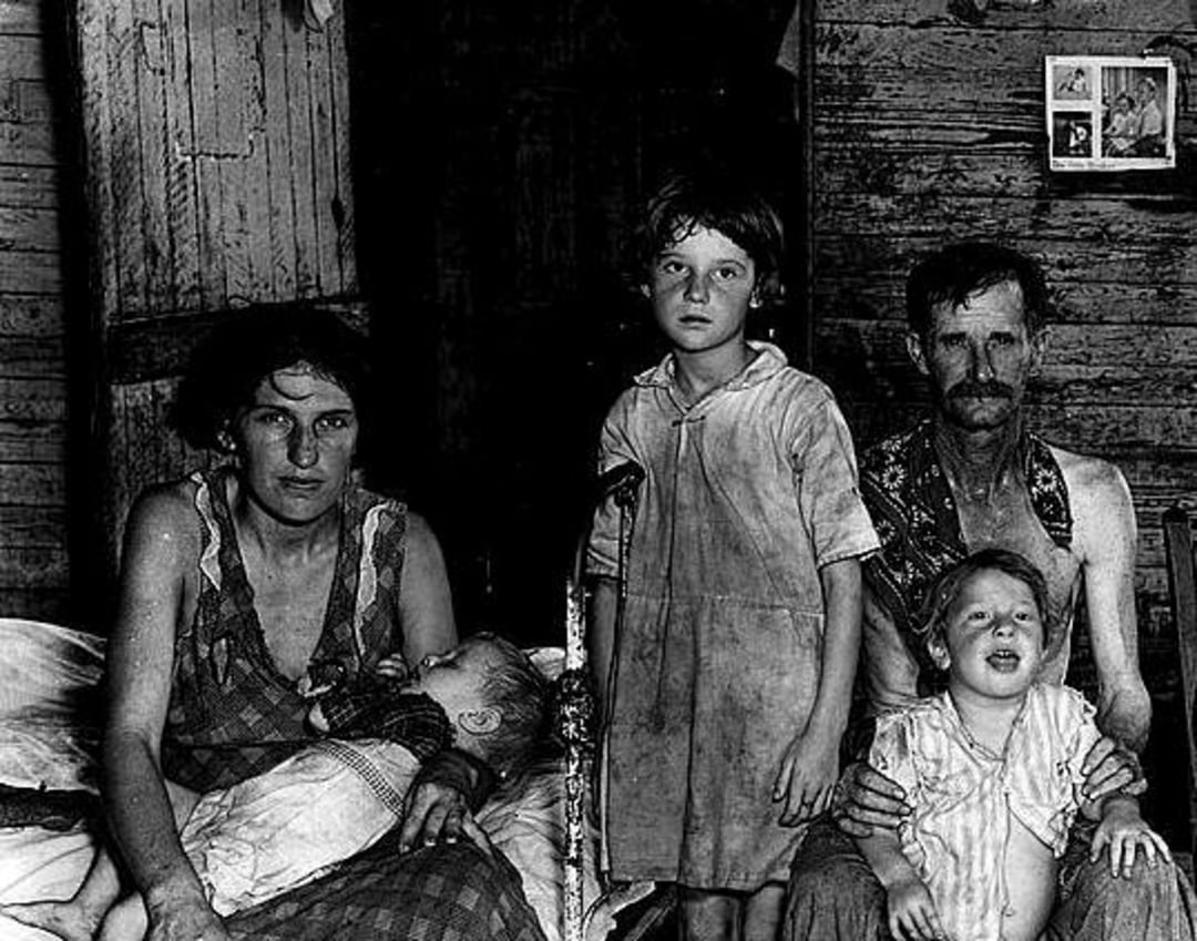 A black-and-white photo of a family in worn clothes inside a rustic wooden room; a woman holds a baby, a man and two children sit and stand nearby, all with serious or tired expressions.