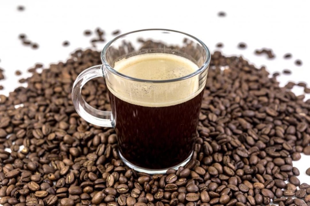 A clear glass mug filled with black coffee sits on a surface surrounded by a pile of roasted coffee beans against a white background.