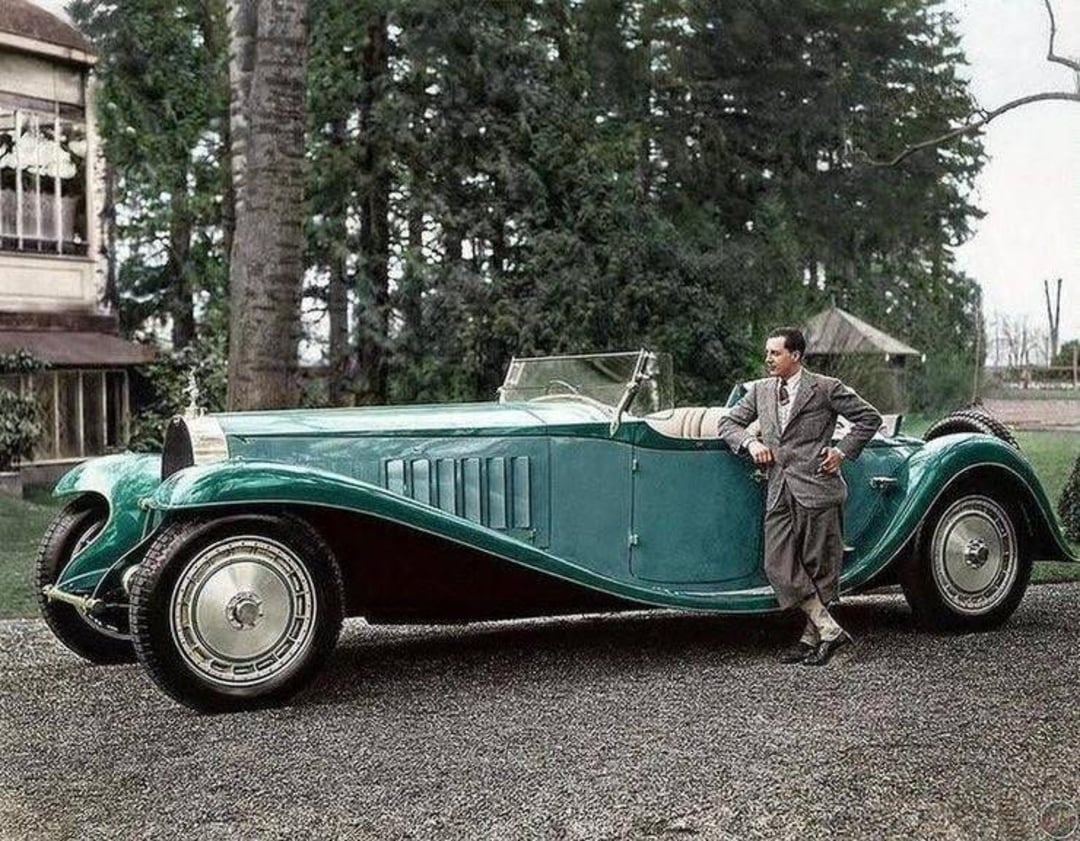 A man in a suit leans against a vintage teal convertible car parked on a gravel driveway, surrounded by tall trees and buildings in the background.