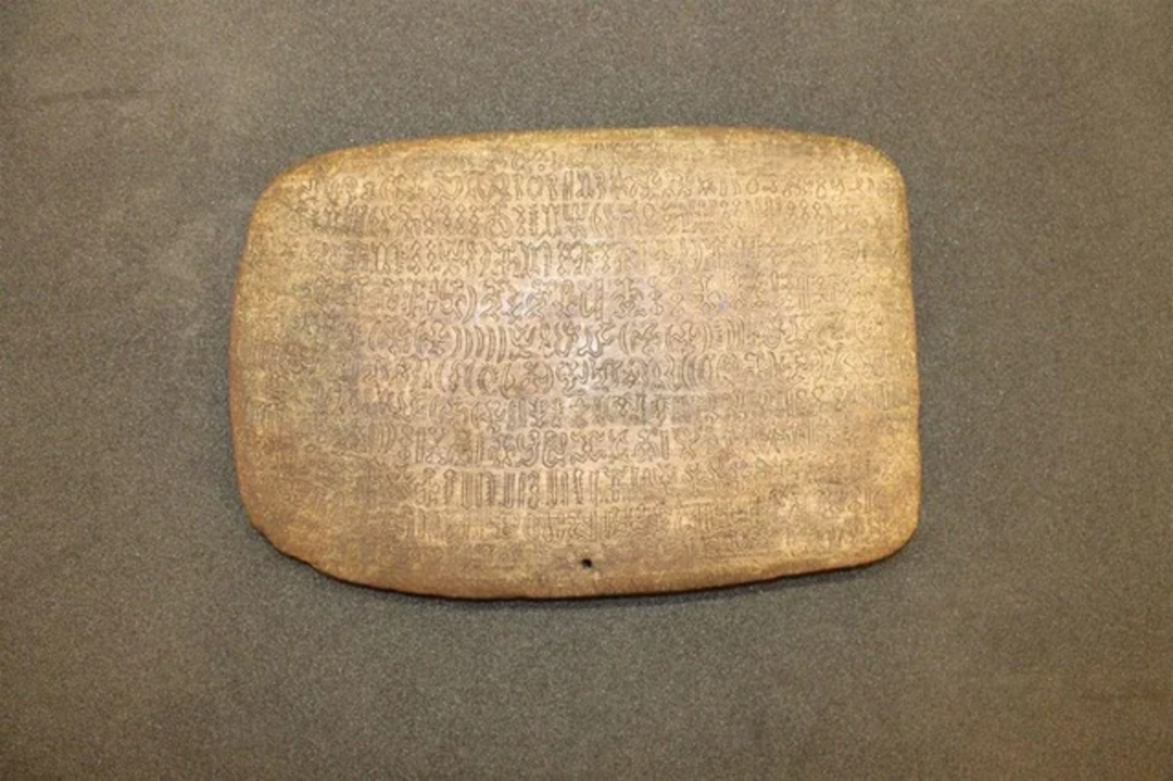 A rectangular, weathered wooden tablet with several lines of faded, intricate script engraved on its surface, displayed on a plain dark background.