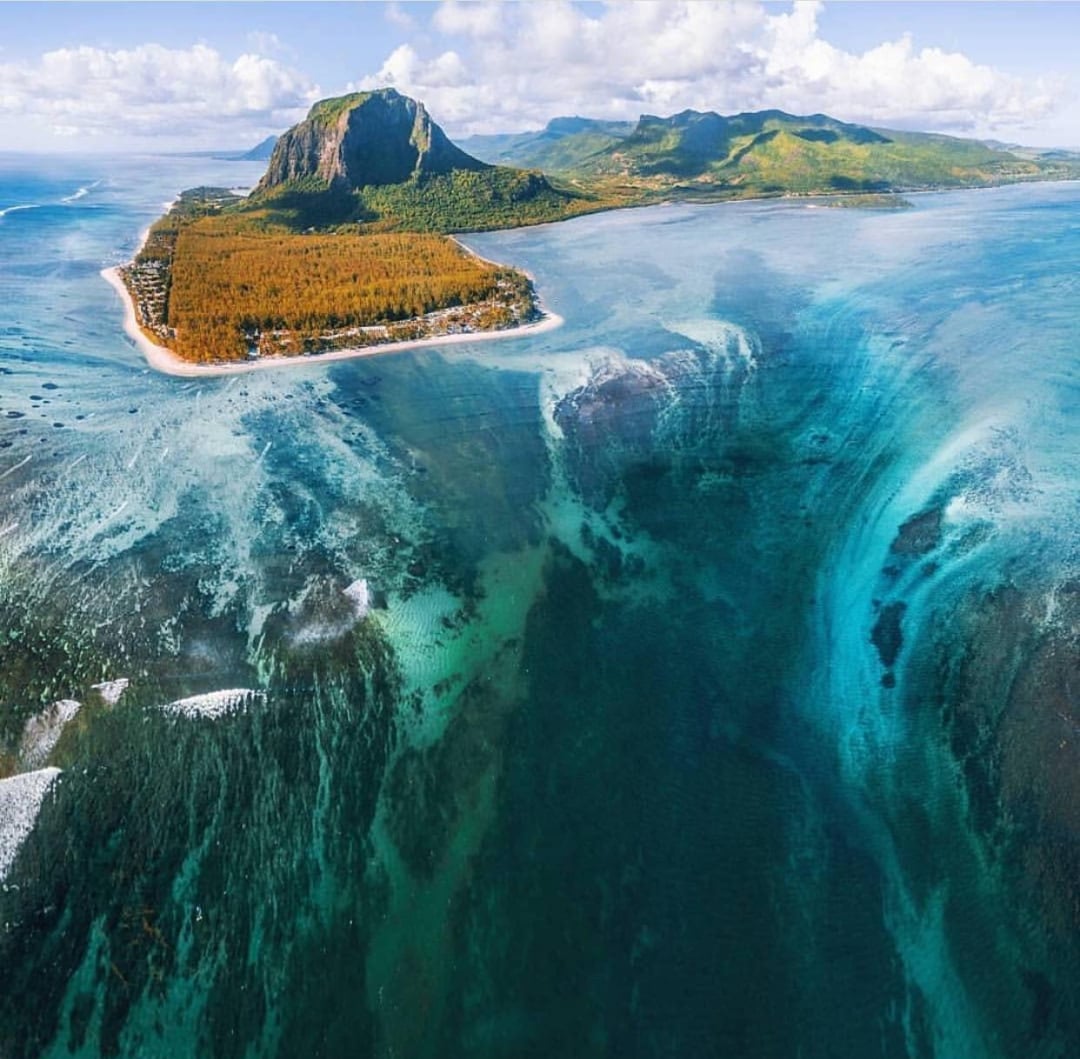 Aerial view of a tropical island with lush greenery, a prominent mountain, and turquoise waters. An underwater waterfall illusion appears near the shore, created by sand and silt patterns beneath the ocean’s surface.