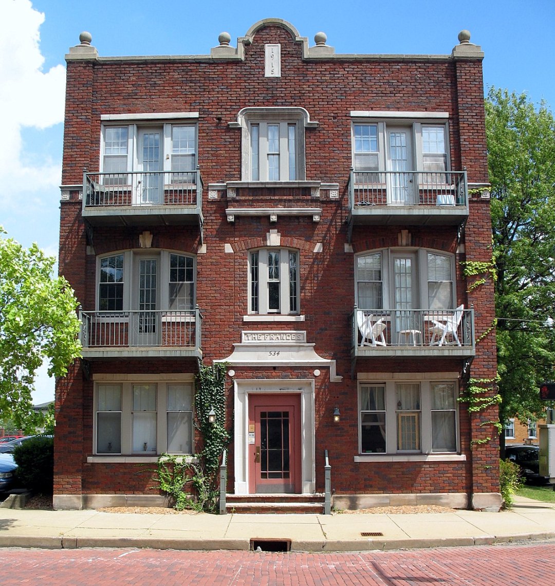 A three-story red brick apartment building with balconies, white trim, and a central entrance labeled “The Spence.” Ivy climbs part of the facade, and trees stand on each side. The street in front is paved with red bricks.