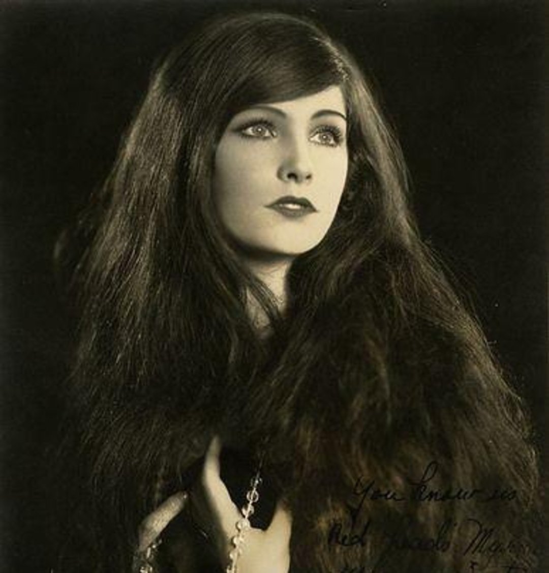 A woman with long, dark hair and striking eyes gazes thoughtfully to the side. She wears a dark fur garment and holds a necklace in her hand. The photo is in black and white, resembling a vintage portrait.