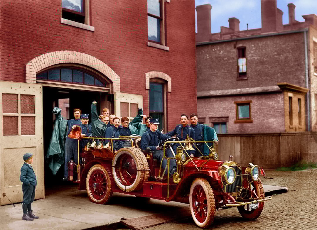 A vintage fire truck with firefighters in uniform is parked outside a brick fire station. Some firefighters wave and smile while a boy stands nearby watching. The scene appears to be from the early 20th century.