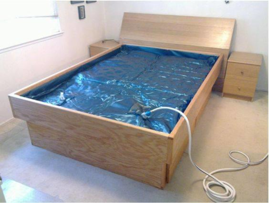 A wooden frame waterbed with a blue vinyl mattress partially filled, a white hose attached for filling, and two bedside tables in a simple, carpeted bedroom.