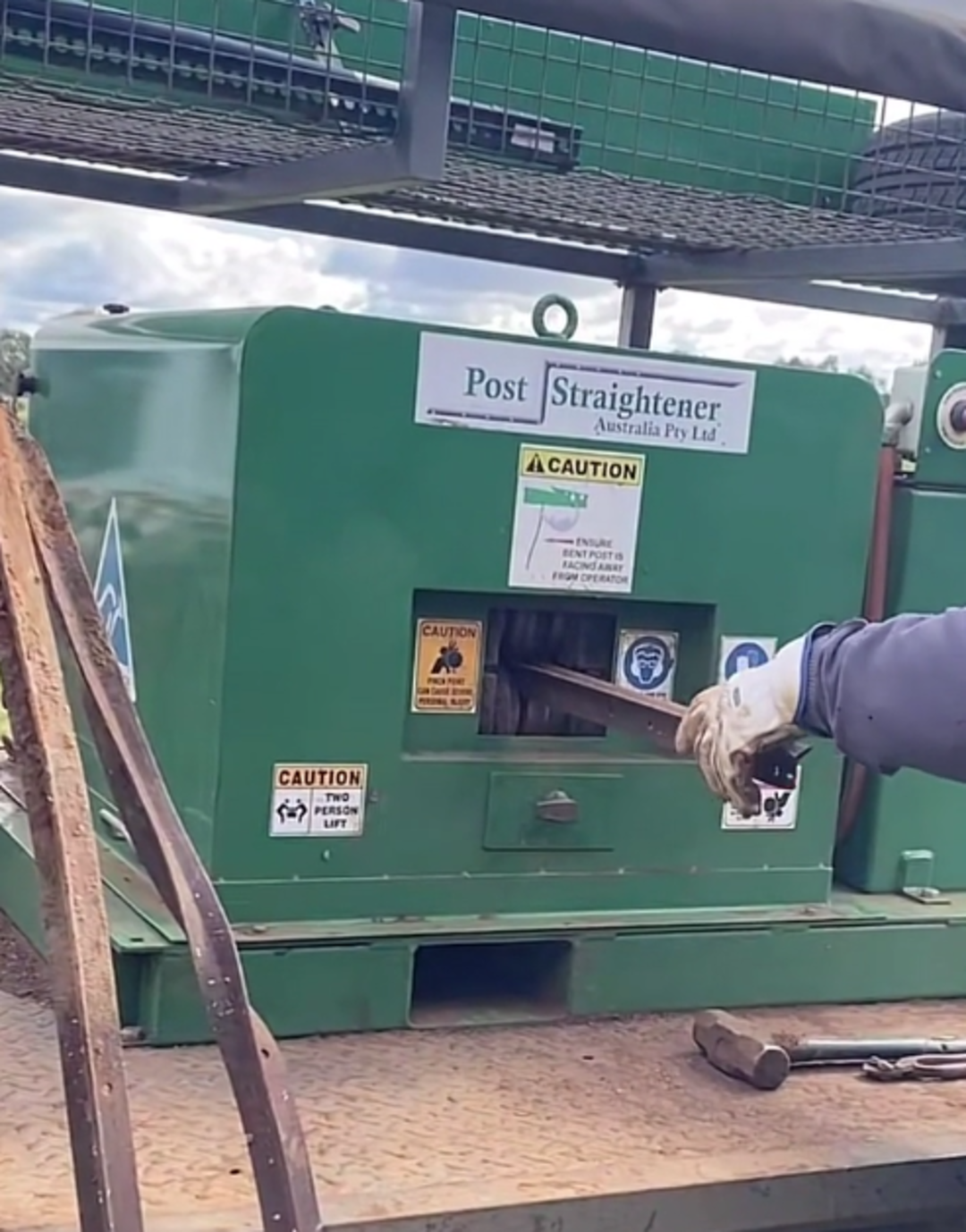 A person wearing gloves inserts a bent metal post into a green post straightener machine labeled with warning signs and instructions, positioned on a metal surface outdoors.