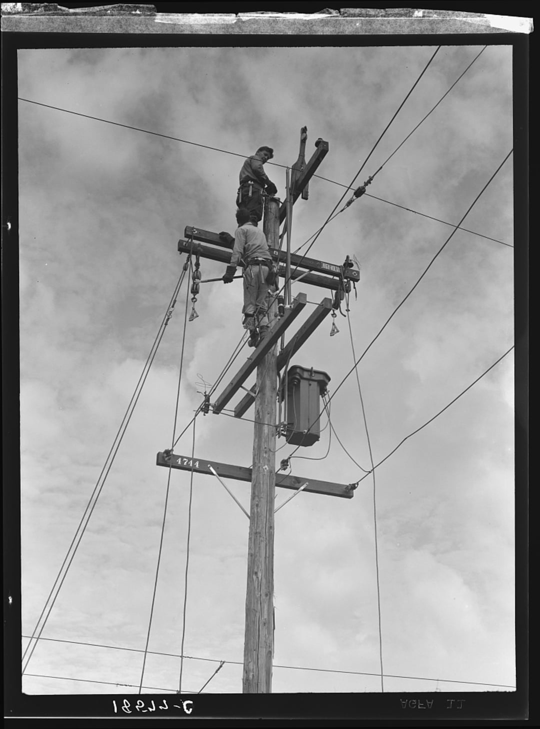 Two utility workers stand on a wooden power pole, working on electrical wires and equipment against a cloudy sky. One climbs near the top, while the other is positioned slightly lower.