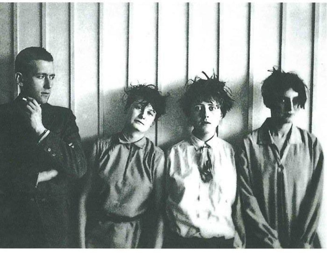 Four people stand in a row against a paneled wall; a man with crossed arms stands beside three women with disheveled hair and expressions ranging from blank to anxious. The photo is black and white and appears vintage.