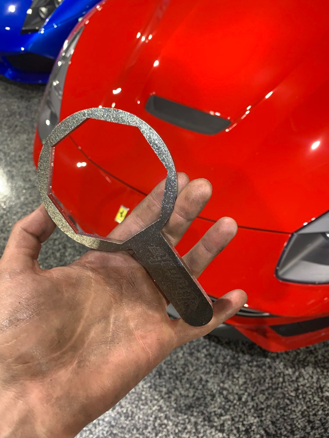 A dirty hand holds a large, metal, octagonal wrench in front of a shiny red sports car with a Ferrari emblem, parked on a speckled garage floor.