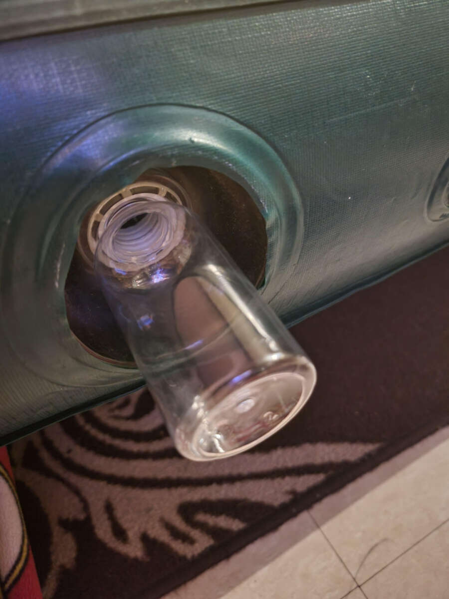 A clear glass tumbler is inserted upside down into the opening of a green water storage tank, with part of the tank and patterned floor mat visible.