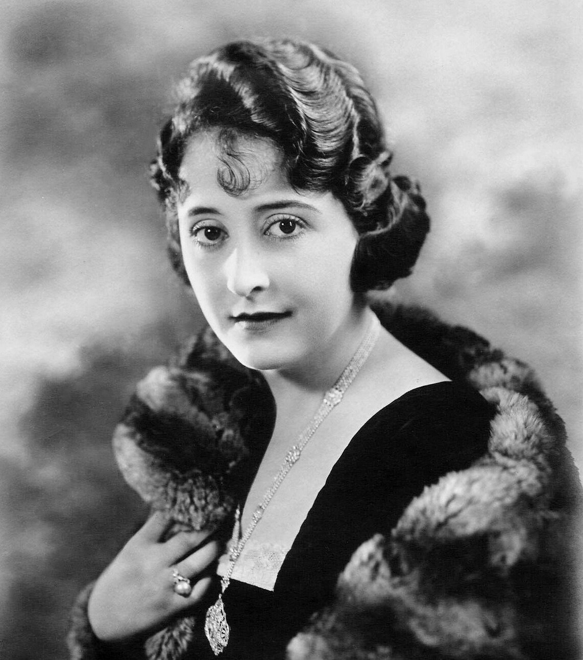 A woman with wavy, short dark hair poses in a formal studio portrait. She wears a fur stole, a dark dress, a necklace, and a ring, and looks directly at the camera with a calm expression. The background is softly blurred.