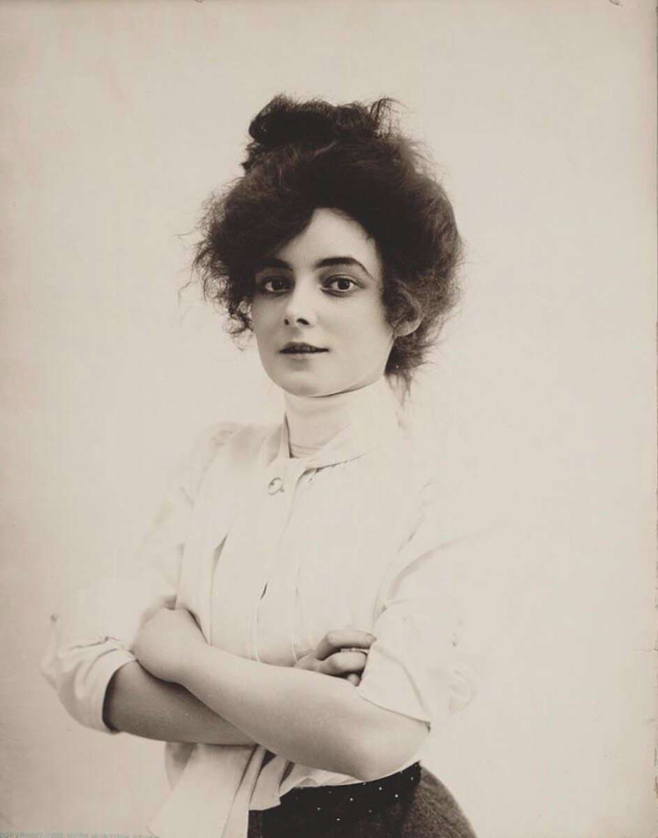 A young woman with dark, wavy hair styled up poses against a plain background. She wears a high-collared white blouse with sleeves rolled up and has her arms crossed, gazing confidently at the camera.