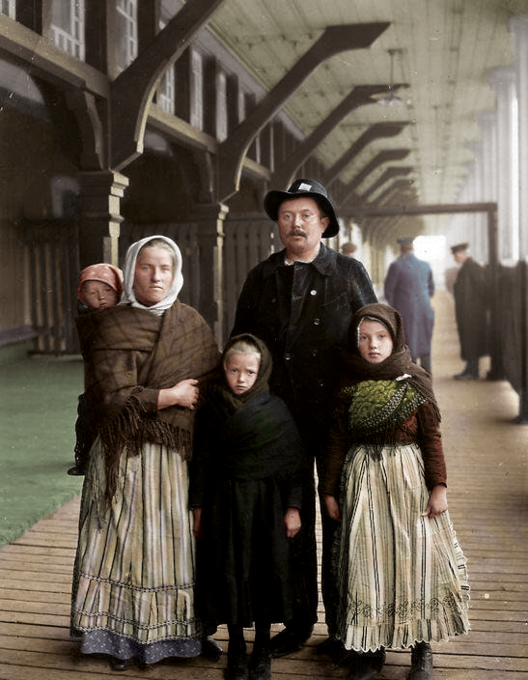 A family of five, including three young girls and their parents, stands on a covered wooden walkway. The adults and children wear old-fashioned, modest clothing and headscarves. The setting appears historical, possibly from the early 1900s.