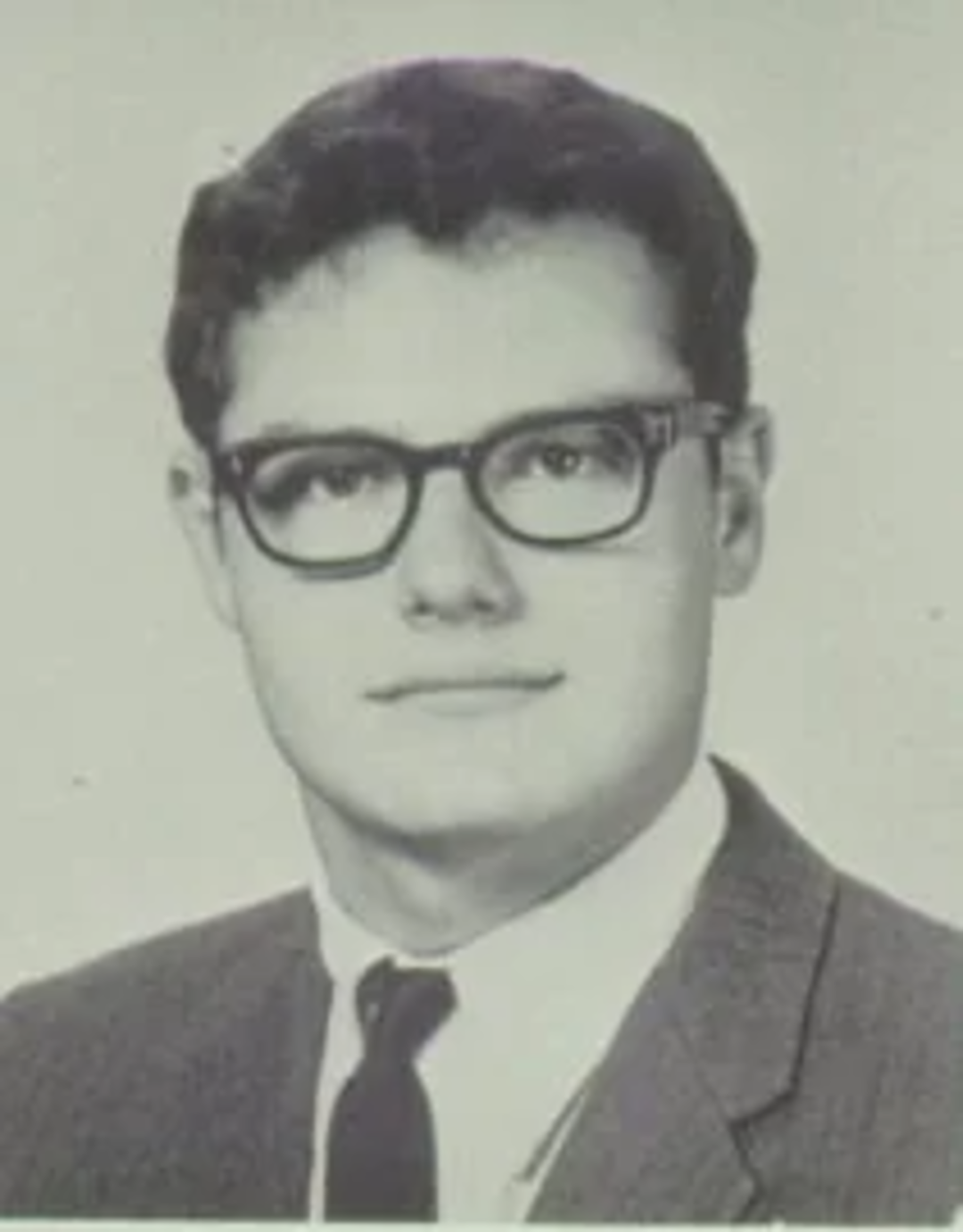 Black-and-white portrait of a young man wearing glasses, a suit, a white shirt, and a tie, with short dark hair and a neutral expression, against a plain background.