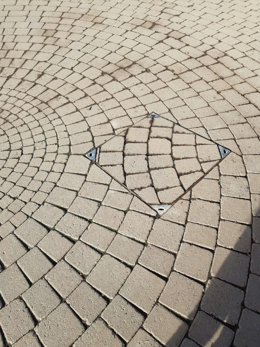 A paved surface with circular patterns of beige bricks, featuring a central square section rotated 45 degrees, disrupting the pattern. Four metal hinges secure the square, likely an access hatch. Shadows appear at the bottom right.