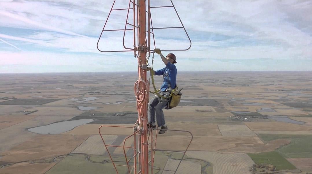 A worker wearing safety gear climbs a tall communication tower high above a flat, rural landscape with fields and ponds visible in the distance under a cloudy sky.
