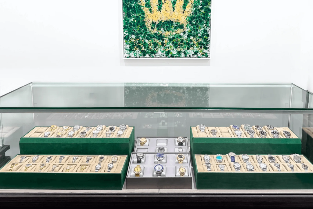 A glass display case full of luxury watches, arranged in rows on green-lined trays, sits beneath a framed artwork featuring a gold crown on a green and white background.