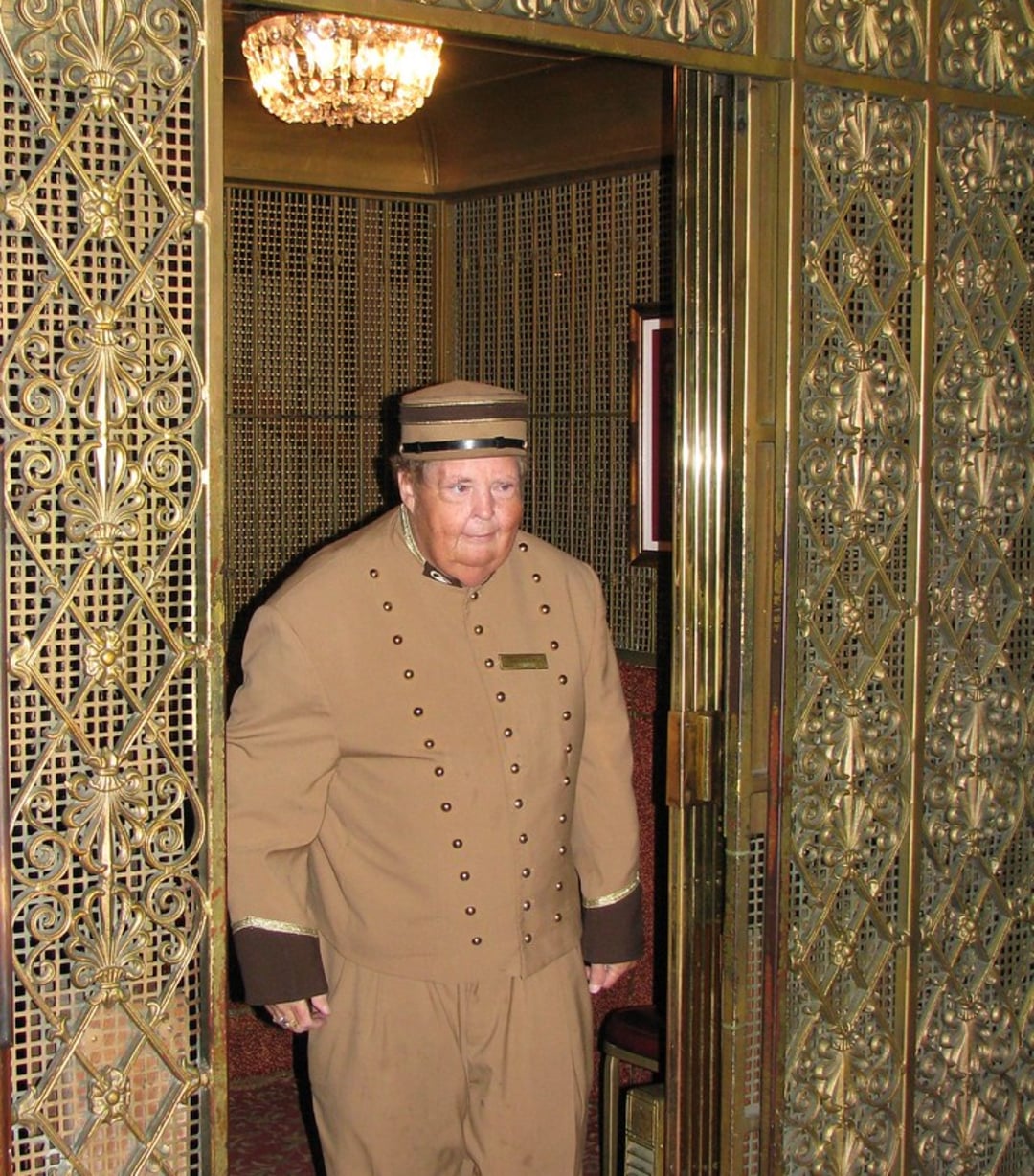An older elevator operator in a brown uniform and hat stands inside an ornate, gold-colored elevator with decorative metalwork and an open door. A chandelier hangs from the ceiling above him.