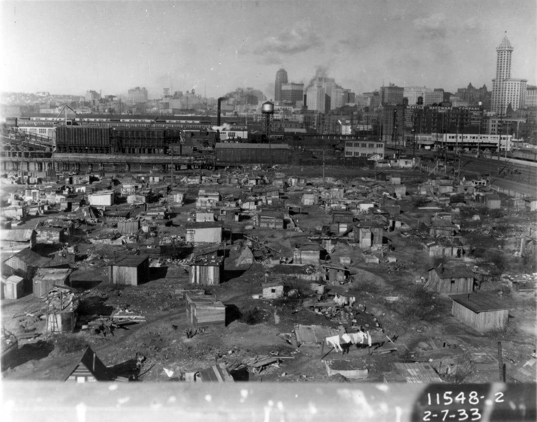 A black-and-white photo shows a large shantytown with many makeshift shelters in a city. Downtown buildings and a clock tower are visible in the background. The image is dated 2-7-33 in the lower right corner.