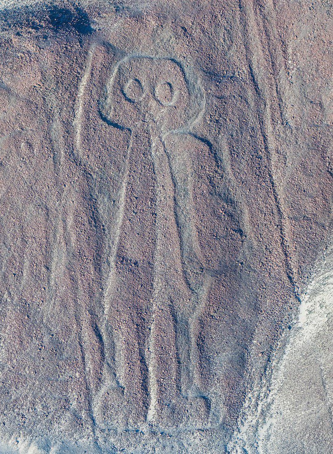 Aerial view of a large geoglyph carved into a rocky surface, depicting a humanoid figure with large round eyes and a raised arm, known as one of the Nazca Lines in Peru.